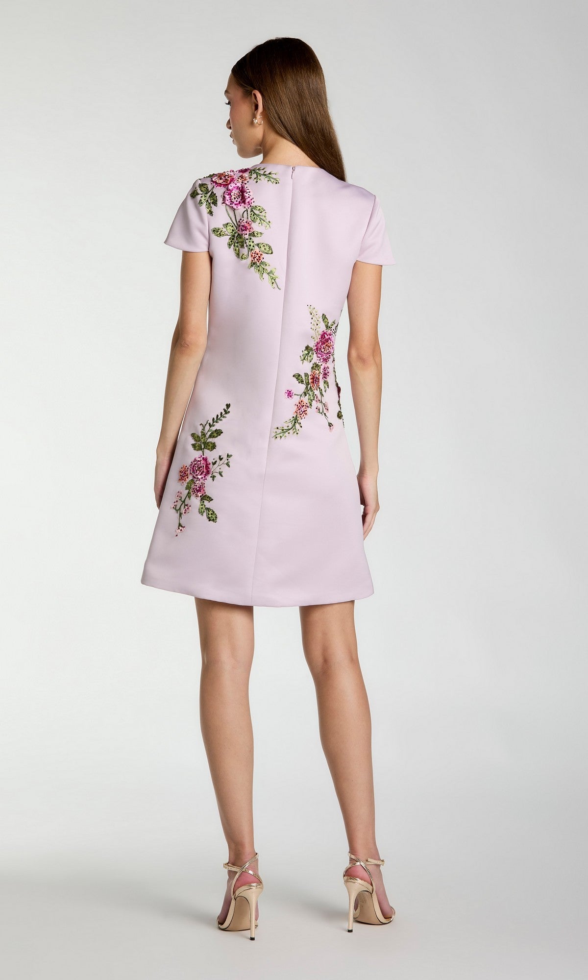 A woman stands facing away, wearing the Mac Duggal 21338 short wedding guest dress—a light pink, short-sleeved design accented with delicate floral embroidery—paired with beige high-heeled sandals against a plain background.